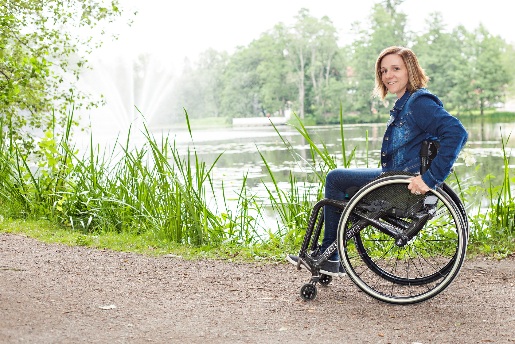 Foto på Josefine Axefall som sitter i rullstolen framför fontänen i Sala Stadspark en solig sommardag. Hon är klädd i blåjeans och jeansjacka.
Fotograf: allprintreklam.se